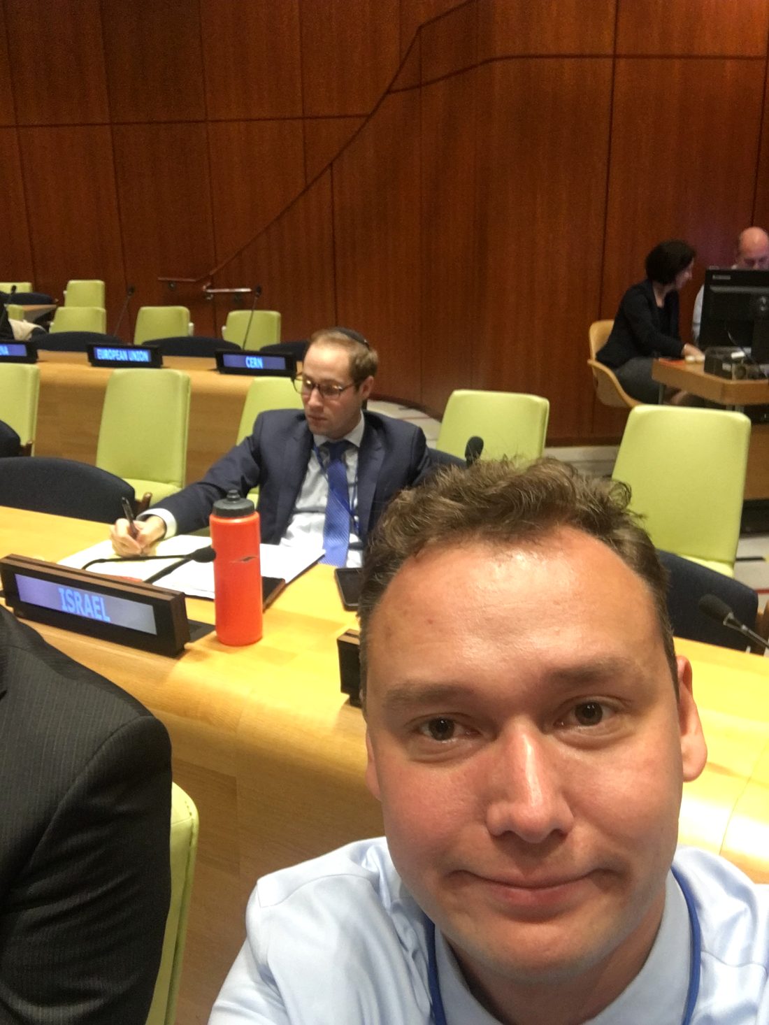 Inside a United Nations committee room — Arseny at the delegate table, with nameplates for Israel, European Union, and CERN visible in the chamber
